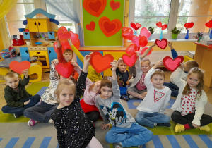 Dzieci siedzą obok siebie na dywanie i podnoszą do góry papierowe serduszka w kolorze czerwonym. W tle tablica z sylwetami serc, kącik domowy, okno. Na parapecie stoją kwiaty i wazony z balonami w kształcie serc.