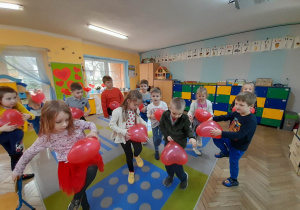 Dzieci tańczą na dywanie z czerwonymi balonami w kształcie serca. W tle szafki z pomocami dydaktycznymi, tablica, okna.