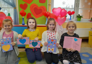 Cztery dziewczynki klęczą na dywanie i prezentują wykonane przez siebie kartki walentynkowe. W tle dekoracja: na tablicy przypięte są czerwone sylwety serc, a po bokach stoją serrduszkowe balony.