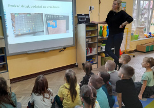 Nauczyciel ze Szkoły Podstawowej nr 6 stoi przy komputerze i tablicy multimedialnej, a dzieci siedzą na dywanie. Pani przedstawia prezentację o szkole.