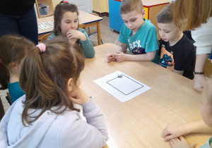 Dzieci wraz z nauczycielem i uczennicą obserwują przy stole poruszanie się ozobota po stworzonej przez siebie planszy.