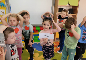 Dzieci z grupy Biedronek stoją trzymając dłonie na swoich głowach. Julka trzyma kartkę z napisem „głowa”