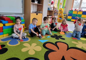 Dzieci z grupy Biedronek siedzą na dywanie, przed nimi leża papierowe figury o różnych kształtach i kolorach