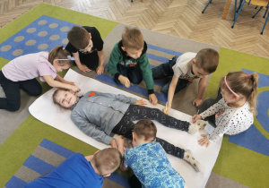 Ala leży na dużym białym arkuszu papieru rozłożonym na dywanie. Pozostałe dzieci z grupy odrysowują jej sylwetkę.