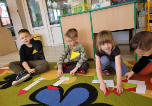 Biedronki siedzą na dywanie. Przed dziećmi leżą figury geometryczne z papieru w różnych kształtach i kolorach, z których dzieci układają różne kompozycje.