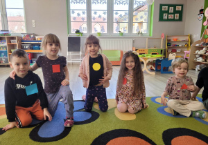 Dzieci z grupy Biedronek siedzą na dywanie. Wszyscy mają przypięte figury w różnych kształtach i kolorach.