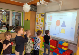 Dzieci z grupy Biedronek stoją przed tablicą multimedialną w tzw. pociąg. Biedronki zaznaczają na tablicy właściwą figurę geometryczną.