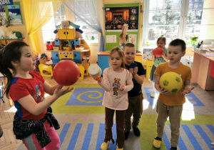 Wiktoria, Vanessa, Bruno, Krzyś i Gabrysia stoją na dywanie w trakcie zabawy ruchowej i trzymają w rękach piłki i garnek w kształcie figury, którą wcześniej pokazał nauczyciel.