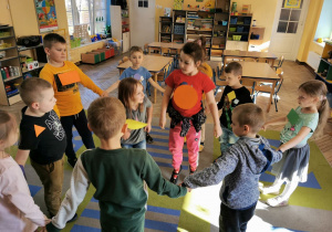 "Słoneczka" stoją w kole na dywanie trzymając się za ręce. Na koszulkach mają przyklejone emblematy z figurami w różnych kolorach. Dzieci z figurami w środku koła poskakują.