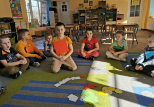 "Słoneczka" siedzą na dywanie. Przed dziećmi na środku leżą figury geometryczne z papieru w różnych kształtach i kolorach.