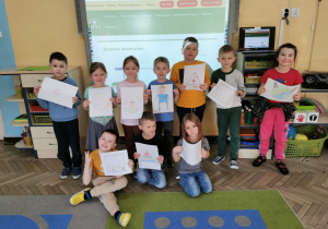 Dzieci z grupy "Słoneczka" stoją przed tablicą multimedialną i prezentują swoje prace - obrazki stworzone na podstawie figury geometrycznej.