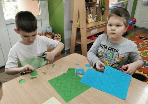 Franek i Artek siedzą przy stoliku i wycinają zielone i niebieskie kółeczka.