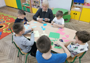 Antoś, Gabryś, Franek, Eryk i Kacper siedzą przy stoliku i wycinają kolorowe kółeczka. Pani z biblioteki pomaga chłopakom w wycinaniu niebieskich kółek.
