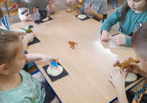 Dzieci siedzą przy stole podczas zabawy "Dino - skamieliny". Wykonują odcisk figurek dinozaurów w masie solnej.