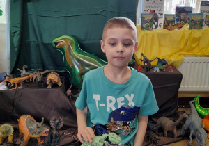 Hubert przykucnął na dywanie na tle dekoracji z okazji Dnia Dinozaura. Chłopiec trzyma szkielet dinozaura.