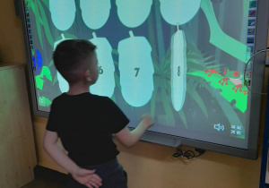 Mieszko stoi przed tablicą multimedialną i szuka pary dinozaurów podczas gry "Memory".