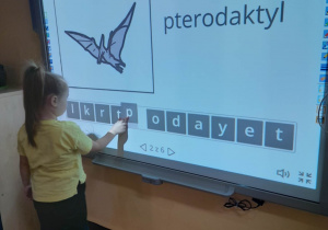 Alicja stoi przed tablicą multimedialną i układa z liter nazwę dinozaura - pterodaktyl.