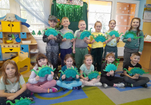 Dzieci pozują do zdjęcia z wykonanymi przez siebie dinozaurami z papierowych talerzyków. W tle dekoracja z okazji Dnia Dinozaura.