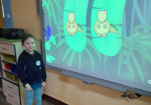 Amelia stoi przed tablica multimedialną. Dziewczynka odkryła parę dinozaurów podczas gry "Memory".