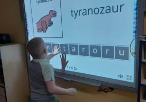 Bruno stoi przed tablicą multimedialną i układa z liter nazwę dinozaura - tyranozaur.