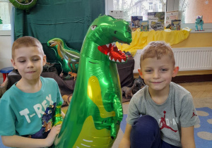 Dwóch chłopców przykucnęło na dywanie, a pomiędzy nimi stoi balon w kształcie dinozaura. W tle dekoracja z okazji Dnia Dinozaura.