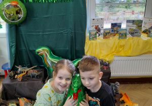Alicja i Mieszko przykucnęli na dywanie, a pomiędzy nimi znajduje się balon foliowy - dinozaur. W tle dekoracja z okazji Dnia Dinozaura.