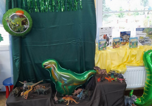 Dekoracja z okazji Dnia Dinozaura. Na zielonym materiale przypiętym do tablicy umieszczony jest napis Dino party oraz dinozaurowy balon foliowy w kształcie koła. Niżej, na brązowym materiale stoją figurki dinozaurów, balon foliowy w kształcie dinozaura, a na parapecie znajduje się wystawa książek o dinozaurach.