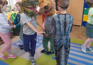 Dzieci tańczą w małych kółeczkach na dywanie przy dźwiękach muzyki. Trzech chłopców ma na sobie maski dinozaurów.