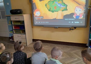Dzieci siedzą na dywanie i ogladąją zaprezentowany na tablicy multimedialnej film o dinozaurach.