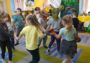 Dzieci tańczą w kole przy piosence o dinozaurze. W srodku troje dzieci w maskach dinozaurów.