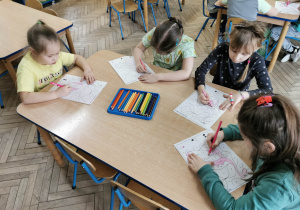 Cztery dziewczynki siedzą przy stole. Na środku leżą kredki na niebieskiej tacce. Dzieci kolorują obrazek dinozaura wg kodu.