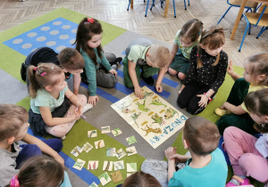 Przedszkolaki siedzą na dywanie, a przed nimi leży plakat przedstawiający różne gatunki dinozaurów. Bruno dokłada element pasujący do wybranego dinozaura.