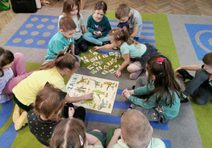 Dzieci siedzą na dywanie, przed nimi leży plakat przedstawiający różne gatunki dinozaurów i pasujące do nich elementy. Alicja dokłada element do obrazka.