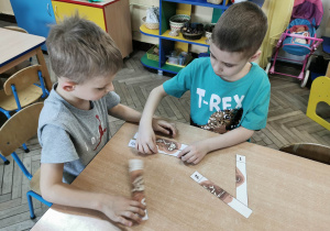 Dwóch chłopców układa przy stole ponumerowane elementy obrazka "Szkielet dinozaura".