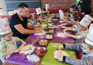 Dzieci z grupy "Słoneczka" siedzą przy stoliku z panem kucharzem i rozwałkowują ciasto według wskazówek.