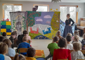Aktorka w roli księżniczki rozmawia z wilkiem (kukiełką). Dzieci siedzą krzesłach i oglądają przedstawienie.