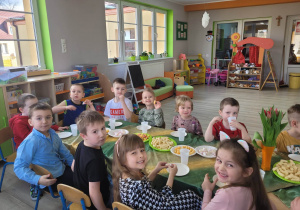 Dzieci z grupy biedronek siedzą przy stole nakrytym zielono - żółtym obrusem. Na stole leżą smakołyki, między innymi pianki, chrupki, ciastka i chipsy.