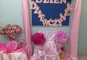 Dekoracja z okazji "Dnia Dziewczynek": na granatowej tablicy przypięty jest napis „Dzień Dziewczynek”, nad tablicą na różowym materiale przypięte są złote korony, przed tablicą stoi krzesełko okryte różowym materiałem, na którym przypięte są różowe kule bibułowe. Z boku na szafce stoją balony w kształcie korony, różowe szarfy, korony oraz różowe pudełeczka.