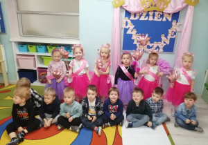 Zdjęcie grupowe dzieci: dziewczynki stoją i trzymają różowe balony w kształcie korony, a chłopcy siedzą po turecku. W tle dekoracja z okazji "Dnia Dziewczynek".