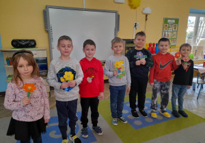 Chłopcy stoją obok siebie i trzymają w rękach własnoręcznie wykonane kwiatki dla dziewczynek.