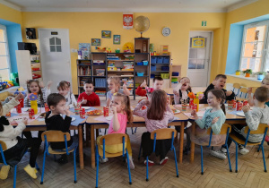 Dzieci siedzą przy połączonych stołach, na których znajduje się słodki poczęstunek. Przedszkolaki z radością podnoszą kciuki do góry.