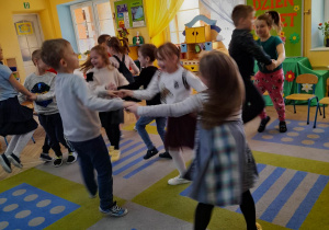 Dziewczynki razem z chłopcami wesoło tańczą na dywanie w małych kółeczkach.