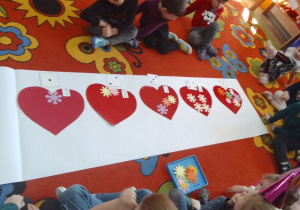 Dzieci siedzą wokół czerwonych serduszek, na których ułożone są kwiatki w ilości, którą wskazują cyfry i kropki na kartonikach.