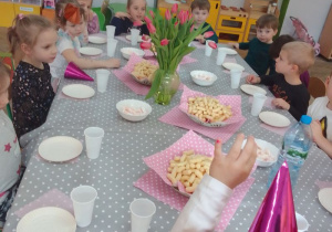 "Motylki" siedzą przy stole, na którym są: wazon z czerwonymi tulipanami, talerze z chrupkami i piankami, papierowe talerzyki i kubeczki.