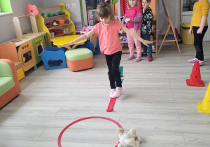 Zosia z grupy Biedronek porusza się po kolorowych szarfach. Przed dziewczynką widać inny fragment toru przeszkód składający się z hula hop i pluszowych zabawek leżących po prawej i po lewej stronie