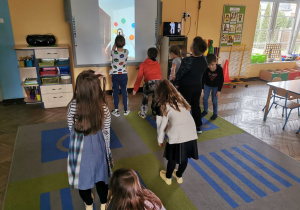 Dzieci z grupy "Słoneczka" w zabawie ruchowej z elementami matematyki na dywanie przed tablicą multimedialną wykonują ruchy zgodnie z tym co wyświetla się na tablicy.