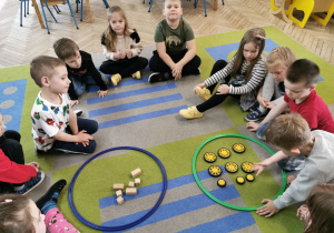 Dzieci z grupy "Słoneczka" siedzą na dywanie, na którym leżą dwie obręcze z posegregowanymi przedmiotami - zabawa "Segregujemy przedmioty".