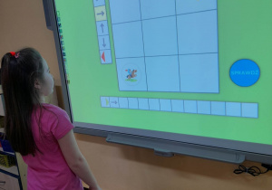 Wiktoria stoi przed tablicą multimedialną podczas zabawy w kodowanie. Dziewczynka wyznacza drogę rycerza do smoka - zapisuje kod za pomocą strzałek.