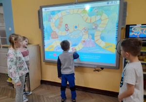 Oliwier przesuwa pionek na planszy podczas gry "Baśniowy las" zaprezentowanej na tablicy multimedialnej. Obok chłopca stoją Alicja, Alicja i Mieszko.