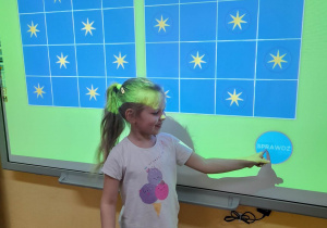 Gabrysia stoi przed tablicą multimedialną, na której wyświetlone jest wykonane przez nią zadanie. Dziewczynka ułożyła gwiazdki na planszy po prawej stronie zgodnie ze wzorem i sprawdza poprawność wykonania zadania.